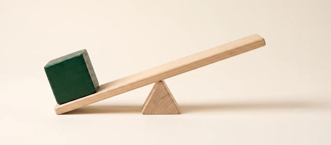 A wooden see-saw tipped hard over: a heavy green weight has pressed one end to the ground, and the raised end is bare.
