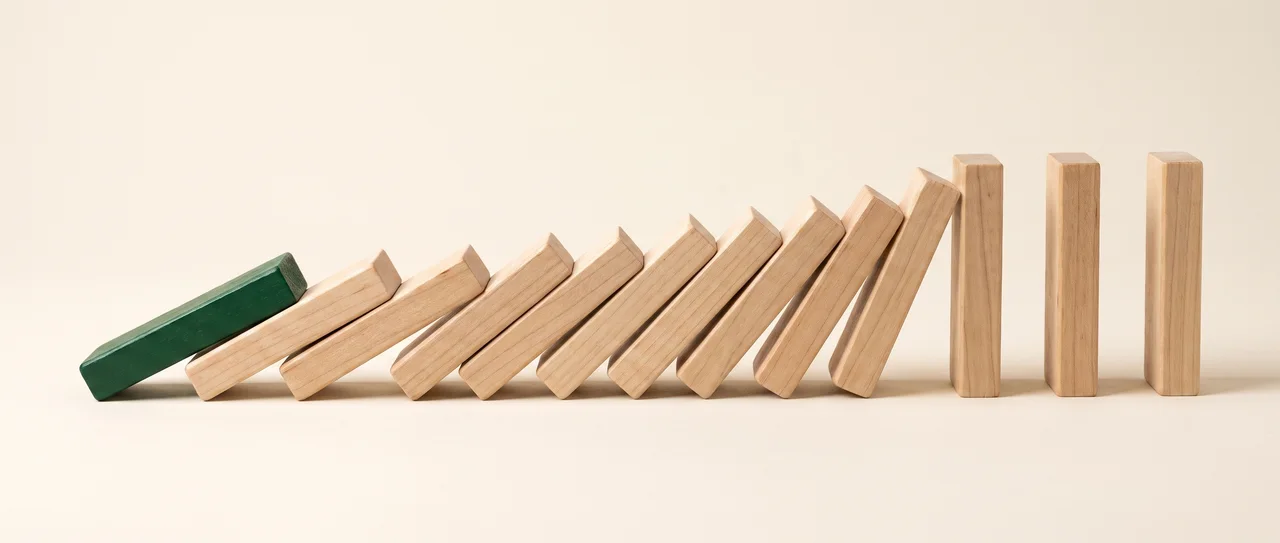 A line of wooden dominoes mid-cascade: one green domino already flat, a run of maple ones falling into each other, and the rest still standing.