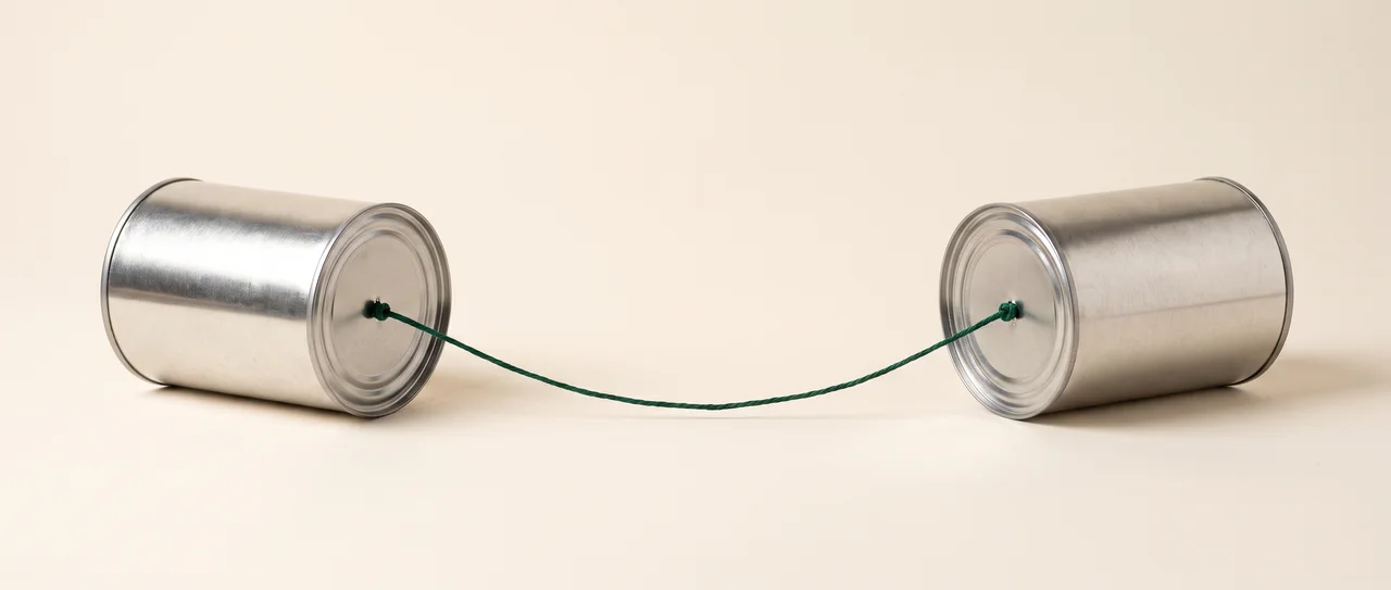 A tin-can telephone: two plain tin cans lying on their sides a long way apart, closed bases facing each other, joined by a single taut green string knotted through the centre of each base.